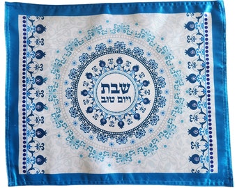 May include: A white and blue decorative mat with a central circular design featuring text in Hebrew. The design includes floral and pomegranate motifs in shades of blue and white, framed by a blue border.