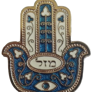 May include: A gold-colored Hamsa hand charm with blue and white enamel detailing. The charm features a blue eye in the center and the Hebrew word "Hamsa" written in white enamel.