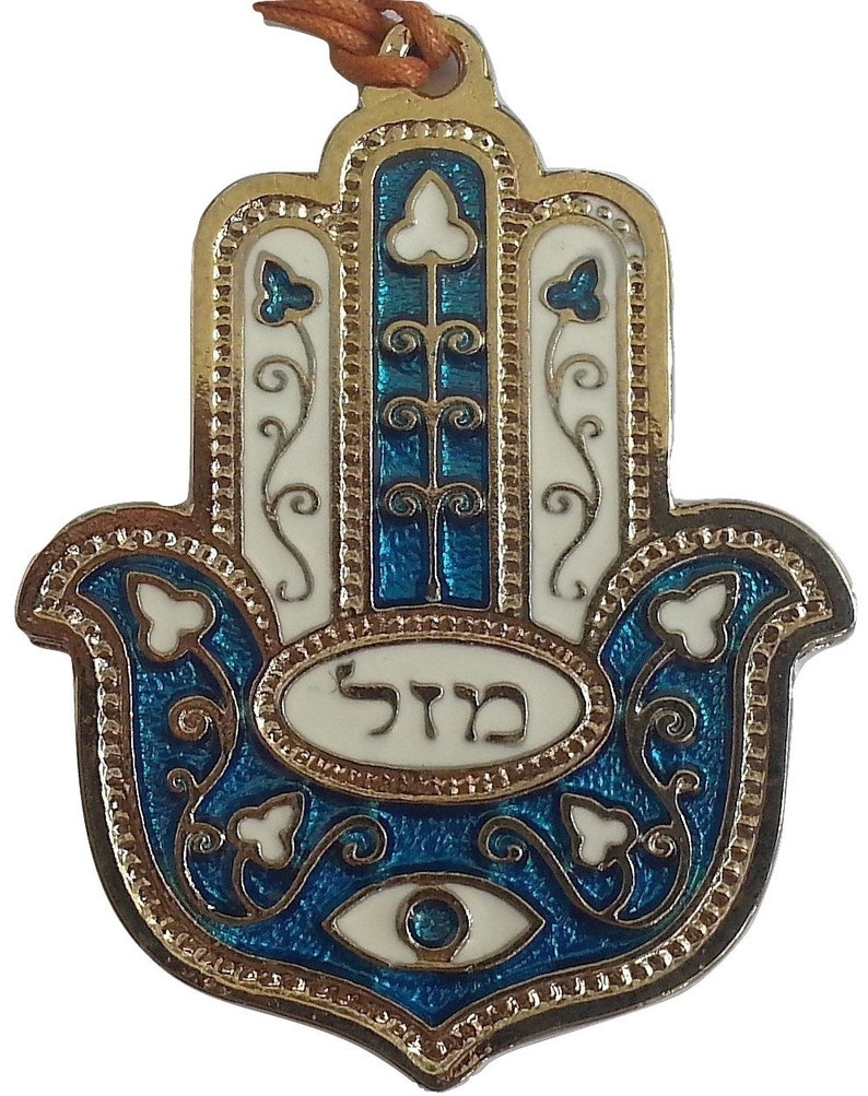 May include: A gold-colored Hamsa hand charm with blue and white enamel detailing. The charm features a blue eye in the center and the Hebrew word "Hamsa" written in white enamel.