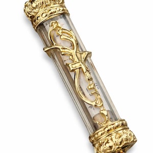 May include: A gold-colored reliquary with a clear, cylindrical body. The reliquary is adorned with ornate gold crown-shaped finials and decorative gold accents. Inside, a scroll and small stones are visible.