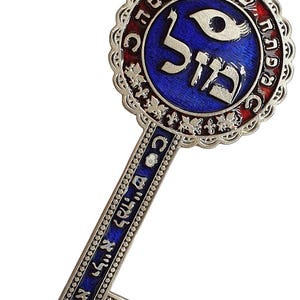 May include: A decorative silver key pendant with blue and red enamel accents. The key features Hebrew text and a blue eye symbol. A brown cord with a turquoise bead is attached for hanging.