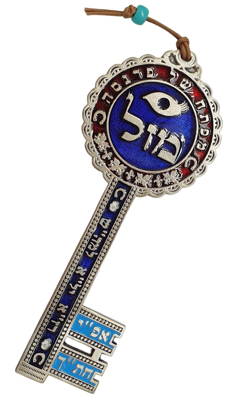 May include: A decorative silver key pendant with blue and red enamel accents. The key features Hebrew text and a blue eye symbol. A brown cord with a turquoise bead is attached for hanging.