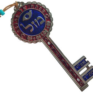 May include: A decorative key pendant with a blue and red design. The key features Hebrew text and a circular emblem. It has a leather cord with a turquoise bead for hanging. The key is made of metal and has intricate details.