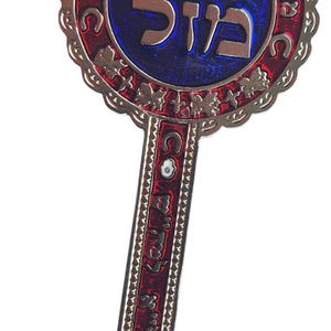 May include: A decorative key pendant with a silver-colored metal frame. The key features red and blue enamel detailing and Hebrew text. A turquoise bead hangs from a brown cord at the top. The key's design includes an eye symbol.