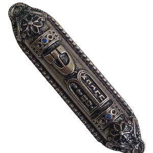 May include: A silver mezuzah with a decorative design and blue accents. The mezuzah has Hebrew writing on it.