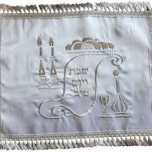 May include: White satin challah cover with silver embroidered details, including a menorah, challah, wine goblet, and Kiddush cup. The Hebrew text "Shehecheyanu" is embroidered in the center.