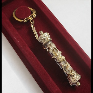 May include: A gold-colored keychain with a clear glass tube containing a scroll-like design. The tube is topped with a crown and the keychain has a ring for attaching to keys.