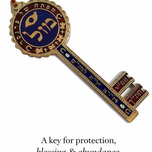 May include: A decorative key with gold-colored metalwork, featuring blue and red enamel accents. The key has Hebrew text and a leather cord with a blue bead. The text on the image reads: "A key for protection, blessing & abundance - always with you."