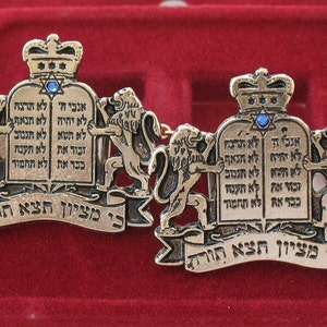May include: Two gold-toned metal cufflinks featuring a design of a crown, two lions, and the Ten Commandments in Hebrew. The cufflinks have a blue stone in the center of the crown and the text "כי מציון תצא תורה" on the bottom.