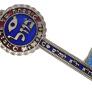 May include: A decorative key pendant with a blue and silver design. The key features Hebrew text and a circular emblem with a blue center. A leather cord with a turquoise bead is attached.