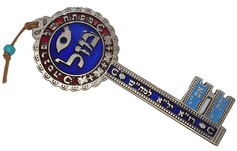 May include: A decorative key pendant with a blue and silver design. The key features Hebrew text and a circular emblem with a blue center. A leather cord with a turquoise bead is attached.