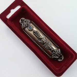 May include: A silver-toned mezuzah case with a blue stone and intricate designs, including a menorah and the hand of God, is displayed in a red velvet case.