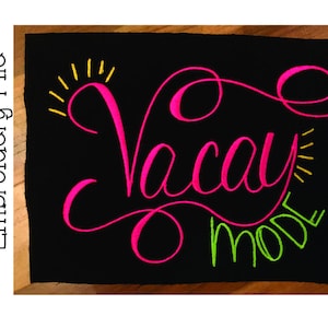 May include: A black fabric with pink and green embroidery that reads "Vacay Mode". The embroidery is in a cursive font with a yellow sunburst above the word "Vacay".