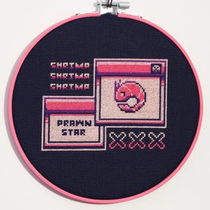 May include: A cross-stitch embroidery on navy blue fabric, set in a pink hoop. The design features pixelated graphics of a shrimp, windows, and the text "PRAWN STAR" and "XXX" in pink and white.