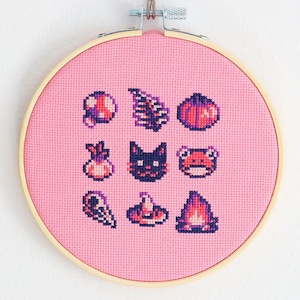 Op de afbeelding: Een roze kruissteek borduurring met negen pixelontwerpen in paars, roze en wit. De ontwerpen omvatten een kat, een kikker en andere grillige vormen. De borduursels zijn geplaatst in een houten ring.