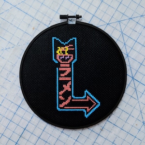 May include: A black embroidery hoop with a pink and blue pixelated arrow pointing to the left. The arrow is outlined in blue and has a yellow and pink pixelated face at the top.
