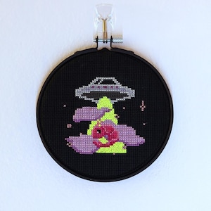 Puede incluir: Un bastidor de punto de cruz negro con un diseño de OVNI. El OVNI es gris, flotando sobre una forma floral verde neón y rosa. El fondo es negro y el bastidor está montado en una pared blanca.