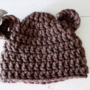 May include: A brown crocheted hat with bear ears. The hat is made with a textured stitch and has a simple design.
