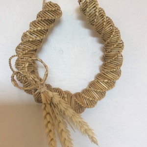 May include: A horseshoe-shaped wreath crafted from woven straw, featuring wheat stalks. The wreath is made of thin, light brown straw pieces, woven together. The wheat adds a natural, rustic touch to the decorative piece.