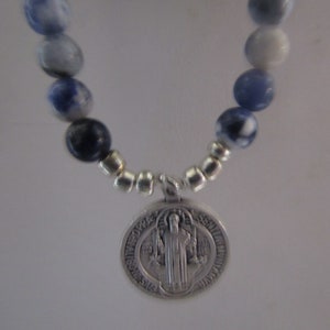 May include: A silver St. Christopher medal charm hangs from a blue and white beaded necklace. The medal features a raised image of St. Christopher carrying a child.