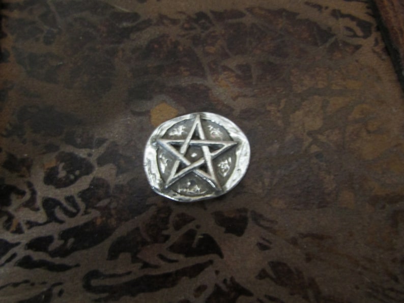 Puede incluir: Un amuleto de pentagrama plateado con un dise&ntilde;o de estrella de cinco puntas. El amuleto es circular y tiene una superficie texturizada.