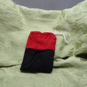 May include: A small, two-toned drawstring bag. The top half is red, and the bottom half is black. A white drawstring is visible at the top. The bag is resting on a light green, patterned fabric.