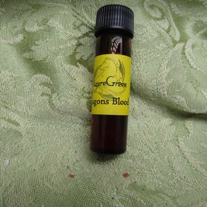 May include: A small, brown glass bottle with a black cap. A yellow label wraps around the bottle, displaying the text "AzureGreen Dragons Blood" with a dragon illustration. The bottle is filled with a dark liquid.