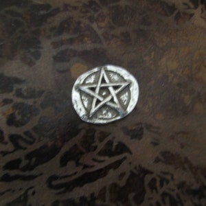 Puede incluir: Un amuleto de metal plateado con un dise&ntilde;o de estrella de cinco puntas. La estrella est&aacute; en relieve y tiene una superficie texturizada.