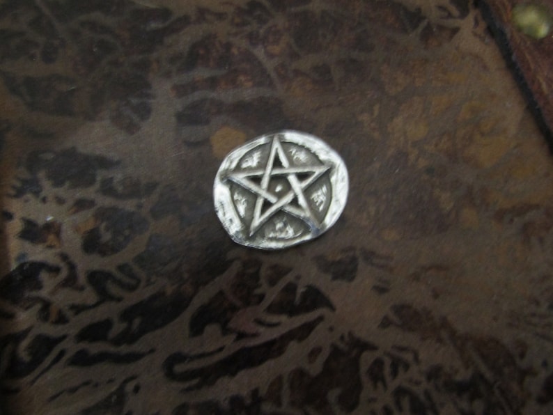 Puede incluir: Un amuleto de metal plateado con un dise&ntilde;o de estrella de cinco puntas. La estrella est&aacute; en relieve y tiene una superficie texturizada.