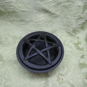 May include: A black, circular cast iron cauldron lid featuring a raised pentagram design. The lid is round with a slightly raised edge and a star symbol in the center. The background is a light green, patterned fabric.