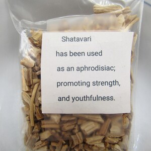 May include: A clear plastic bag filled with dried, light brown Shatavari pieces. A white label on the bag reads: "Shatavari has been used as an aphrodisiac; promoting strength, and youthfulness."