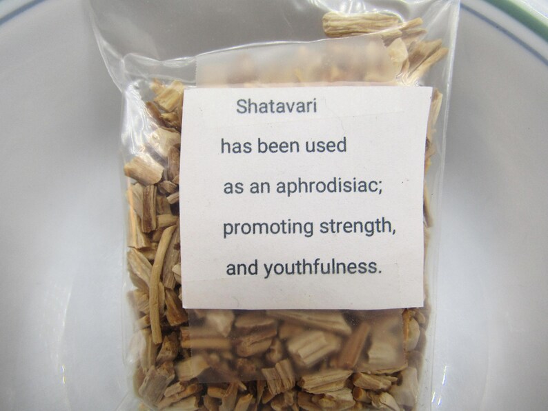 May include: A clear plastic bag filled with dried, light brown Shatavari pieces. A white label on the bag reads: "Shatavari has been used as an aphrodisiac; promoting strength, and youthfulness."