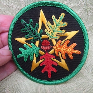 May include: A circular embroidered patch featuring a black background with a central yellow five-pointed star. Four oak leaves in green, yellow, orange, and red are arranged around a small red acorn at the center, all bordered by green stitching.