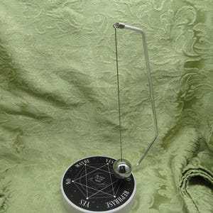 May include: A divination pendulum set with a silver-colored metal frame and a spherical weight. The pendulum hangs over a black and white circular base with the words "YES", "NO", "MAYBE", and "REPHRASE". The background is a green fabric.