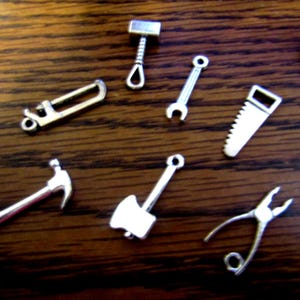May include: A collection of small, silver-colored miniature tools, including a hammer, saw, wrench, and pliers, are arranged on a dark wooden surface. The tools are detailed and appear to be charms or decorative items. The image is well-lit, highlighting the metallic sheen of the tools.