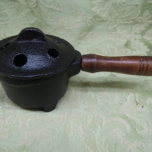 May include: A black cast iron pot with a matching lid featuring three circular vent holes and a central handle. The pot has a short, cylindrical body with a textured surface and a horizontal wooden handle attached to the side for easy gripping.