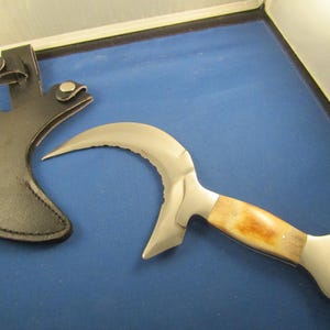 May include: A curved, silver knife with a brown handle and a black leather sheath. The knife has a serrated edge and a pointed tip.