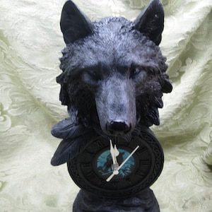 May include: A black wolf head clock with blue eyes and detailed fur. The clock face has Roman numerals and a forest scene. The base features a Celtic knot design. The clock hands are white.