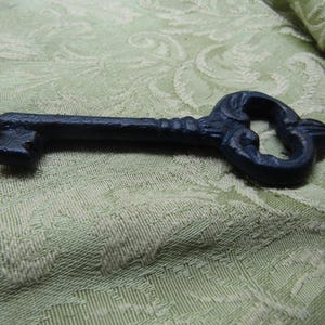 May include: A decorative, dark blue cast iron key with ornate detailing. The key has a classic skeleton key design, with a detailed bow and a textured shaft. The key is approximately 6 inches long.