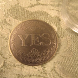 May include: A bronze-colored coin with the word "YES" embossed in the center. The coin has an ornate, decorative border. A clear plastic container is partially visible in the upper right corner. The coin rests on a patterned, light green fabric.