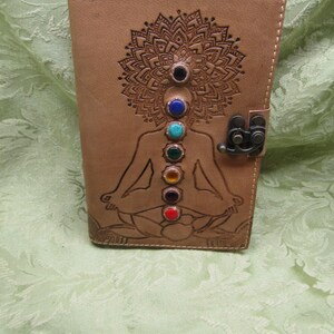 May include: A tan leather journal with a detailed mandala design and a meditating figure etched on the cover. The figure has seven colored gemstones aligned along the body. The journal is closed with a metal clasp.