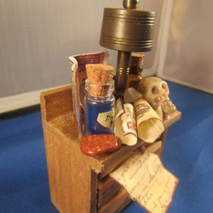 May include: A miniature wooden dresser with three drawers, decorated with a variety of items, including a small lamp, a glass bottle with a cork stopper, a skull, and rolled up papers. The dresser is painted brown and has a natural wood finish.