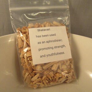 May include: A clear plastic bag filled with light brown, wood-like pieces. A white label on the bag reads "Shatavari has been used as an aphrodisiac; promoting strength, and youthfulness."