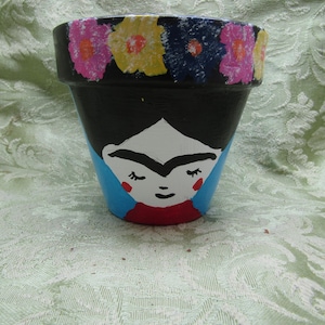 May include: A hand-painted black terracotta flower pot featuring a portrait of a woman with a unibrow, red lips, and a floral design around the rim. The pot has a blue and red painted background.