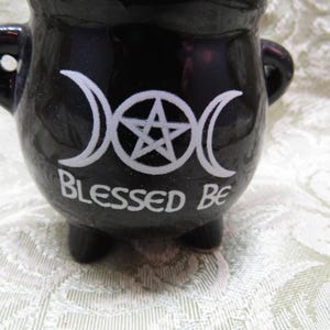 May include: A black ceramic cauldron with three small feet and two handles. The cauldron features a white triple moon symbol and a pentagram, with the words "Blessed Be" in white lettering.