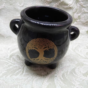 May include: A black ceramic cauldron with a gold tree of life design. The cauldron has two handles and three small feet. The tree of life is a Celtic symbol. The cauldron is likely used for incense or rituals.