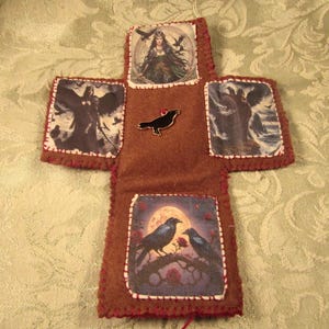 May include: A brown felt cross-shaped item with images of ravens, a woman, and angels. The edges are stitched with red thread. A black raven pin is in the center. The images are printed on fabric and sewn onto the felt.
