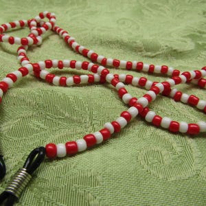 May include: A beaded eyeglass chain with alternating red and white beads. The chain has black rubber loops to attach to glasses and silver metal coils. The chain is laying on a green patterned surface.