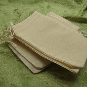 May include: A stack of small, off-white muslin drawstring bags. The bags are rectangular with a stitched seam and a drawstring closure at the top. The bags are stacked on a green patterned fabric.