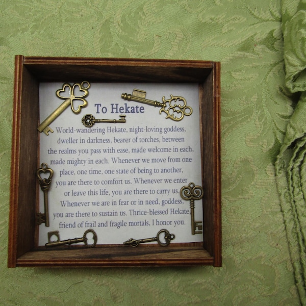 May include: A square wooden box containing a white card with the printed text To Hekate and a prayer, decorated with six antique brass-coloured metal keys of various sizes and designs arranged around the central text on the card surface.